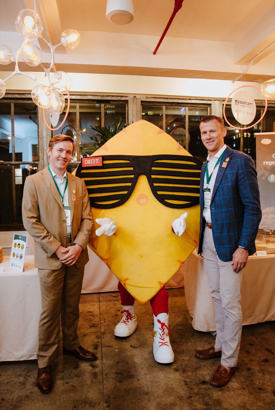 Two people in suits pose beside a mascot shaped like a square cracker wearing sunglasses