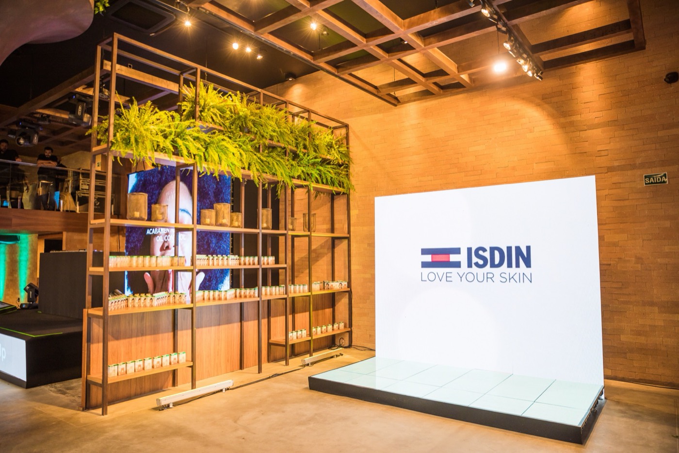 Wooden shelving with plants and products beside a white "ISDIN LOVE YOUR SKIN" panel on a lit platform