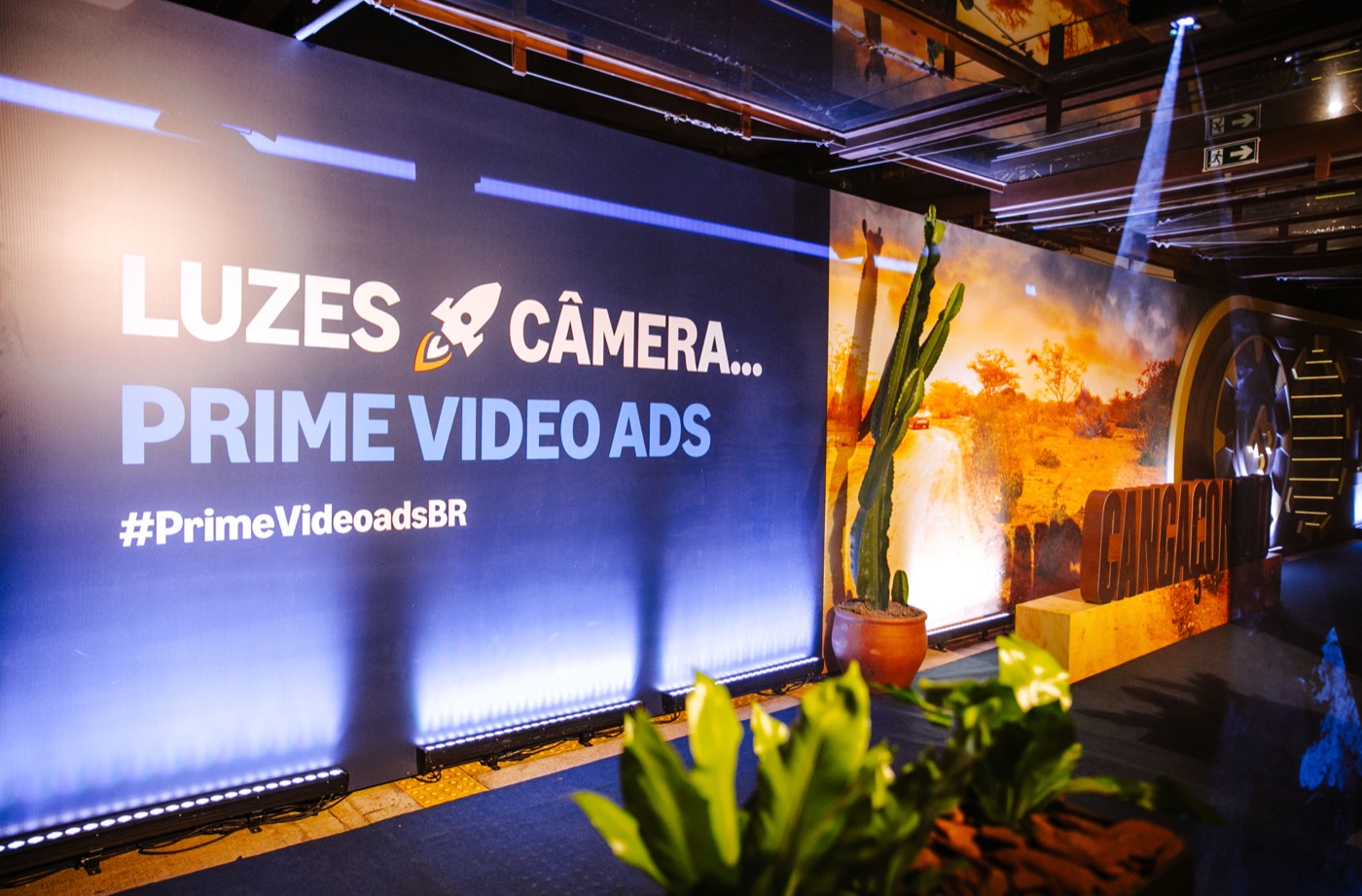 LED panel reading "LUZES CÂMERA... PRIME VIDEO ADS #PrimeVideoadsBR" next to a set with a cactus and plants
