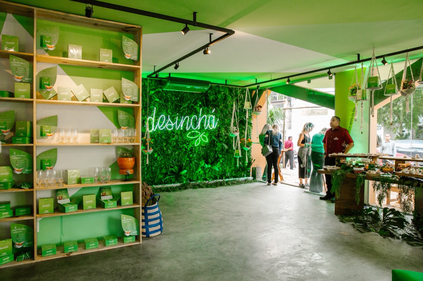 Room with a shelf of green products, a foliage wall with a "desinchá" neon sign and guests by a buffet