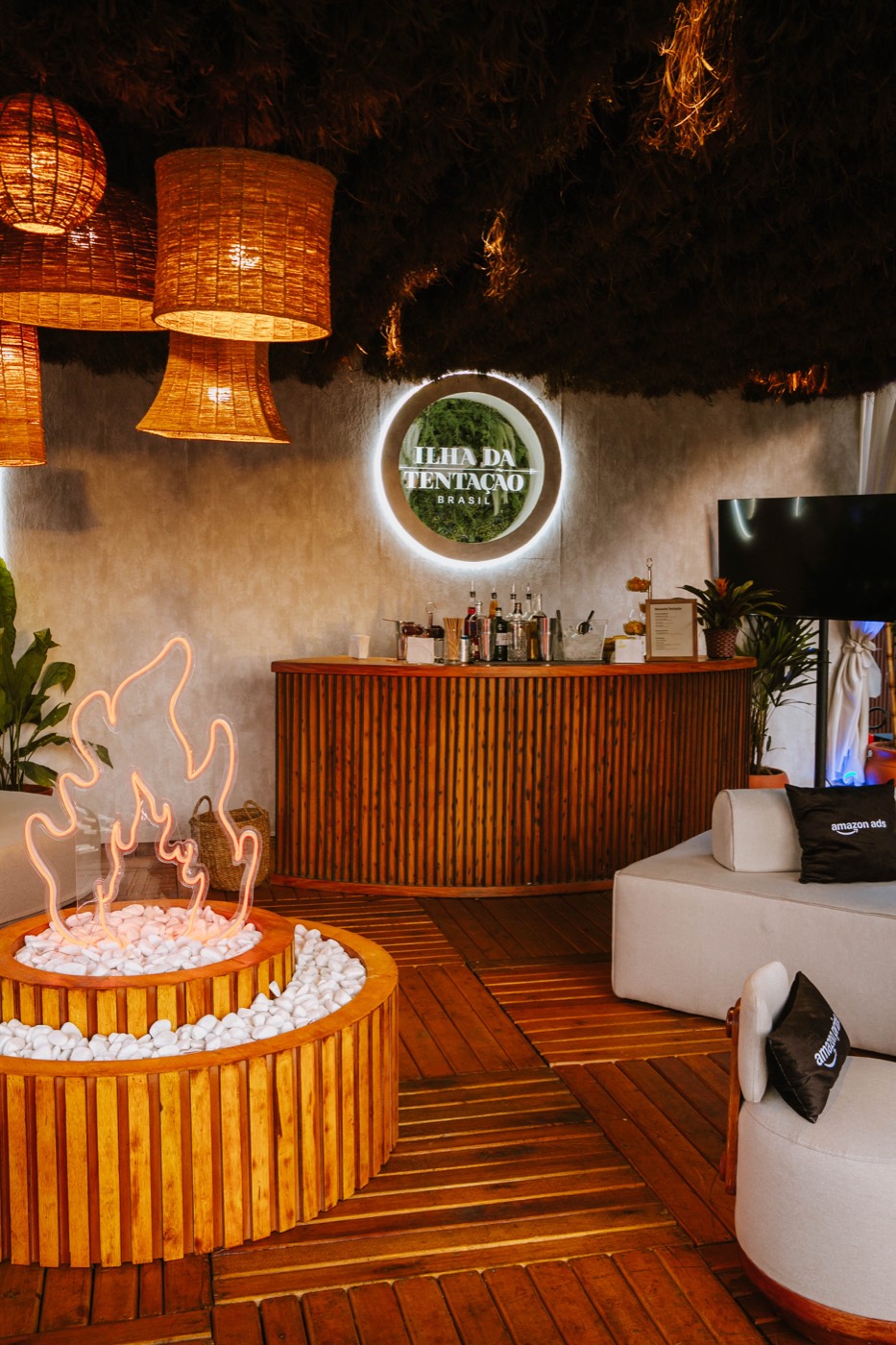 Lounge with a wooden bar, straw pendant lamps, a neon faux campfire and an "ILHA DA TENTAÇÃO BRASIL" sign