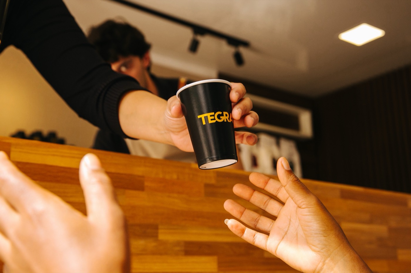 Person behind a wooden counter hands a black cup reading "TEGRA" to outstretched hands in the foreground