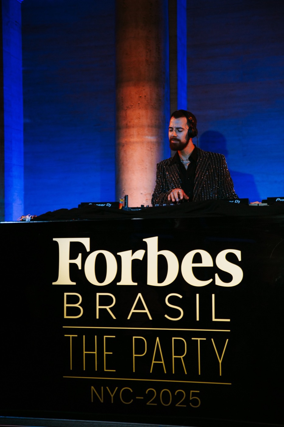 DJ wearing headphones plays behind a black booth with "Forbes BRASIL THE PARTY" lettering, under blue light