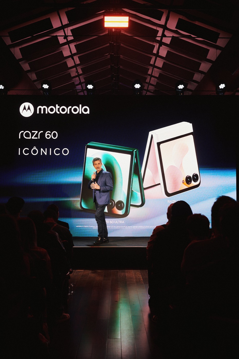 Person on stage before a screen reading "motorola razr 60 ICÔNICO" with images of foldable phones, audience in silhouette