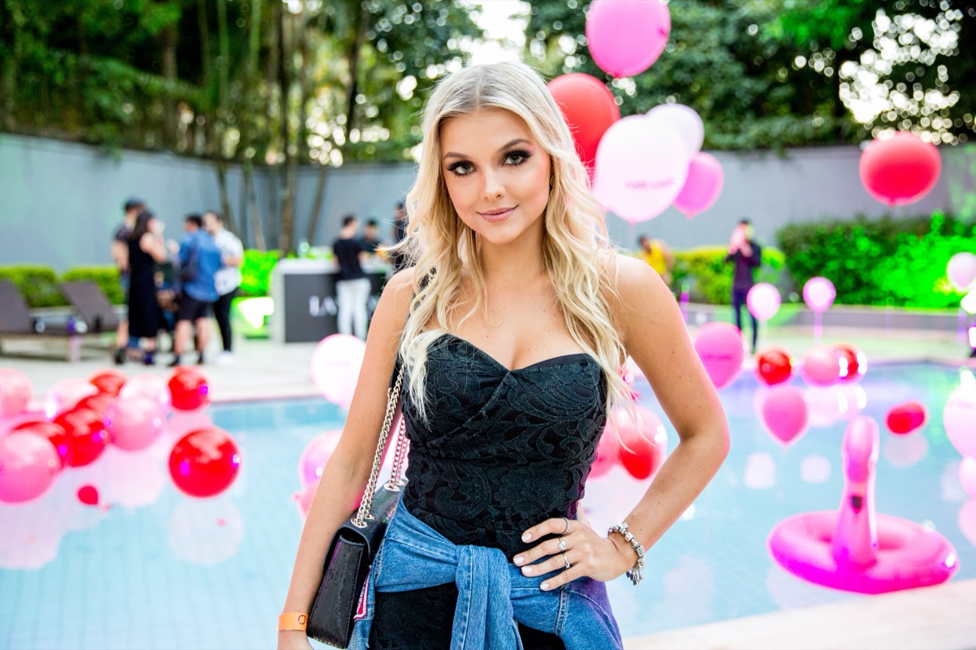 Person with blonde hair poses, hand on hip, in front of a pool with red and pink balloons