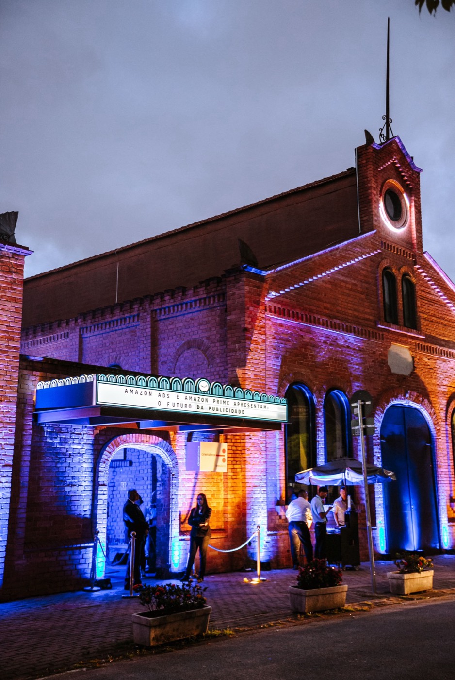 Brick building with a clock tower, a lit marquee and people at the entrance at dusk