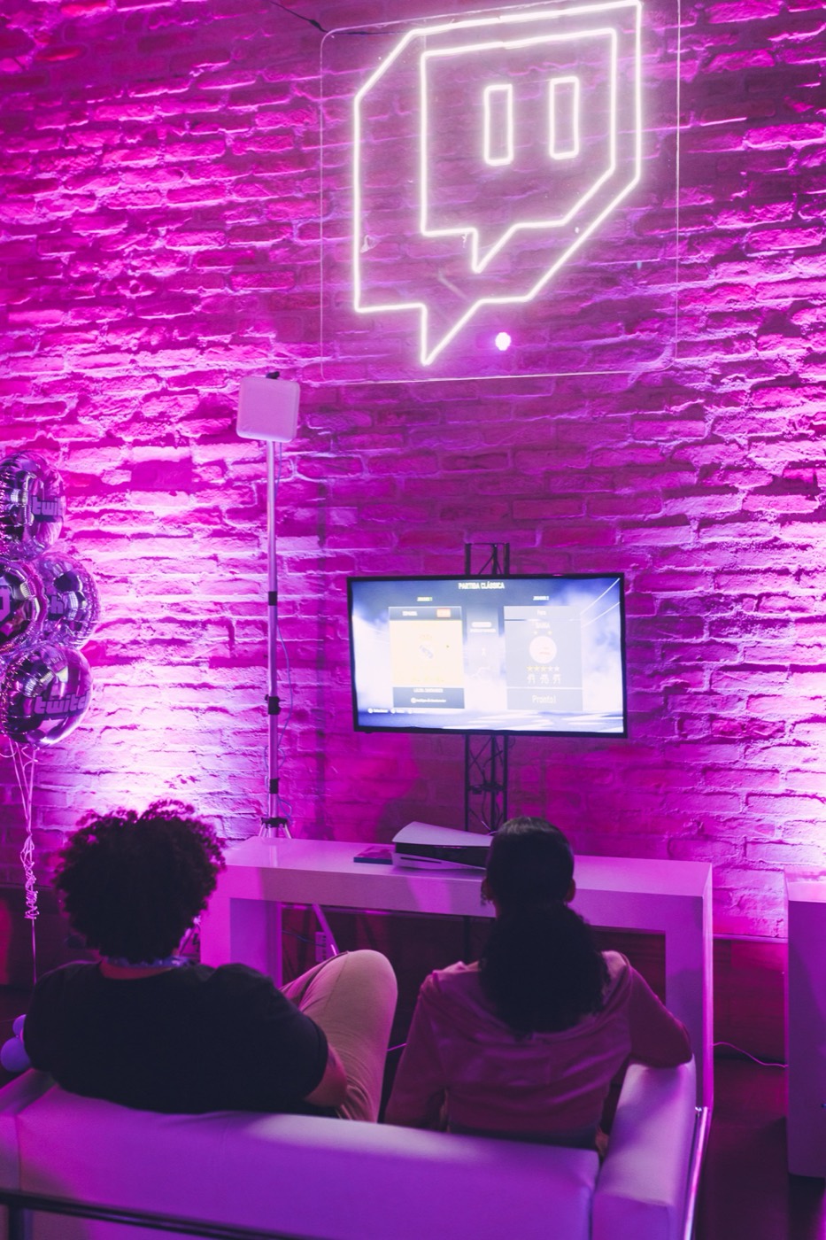 Seen from behind, two people on a white sofa play video games by a pink-lit brick wall with neon logo