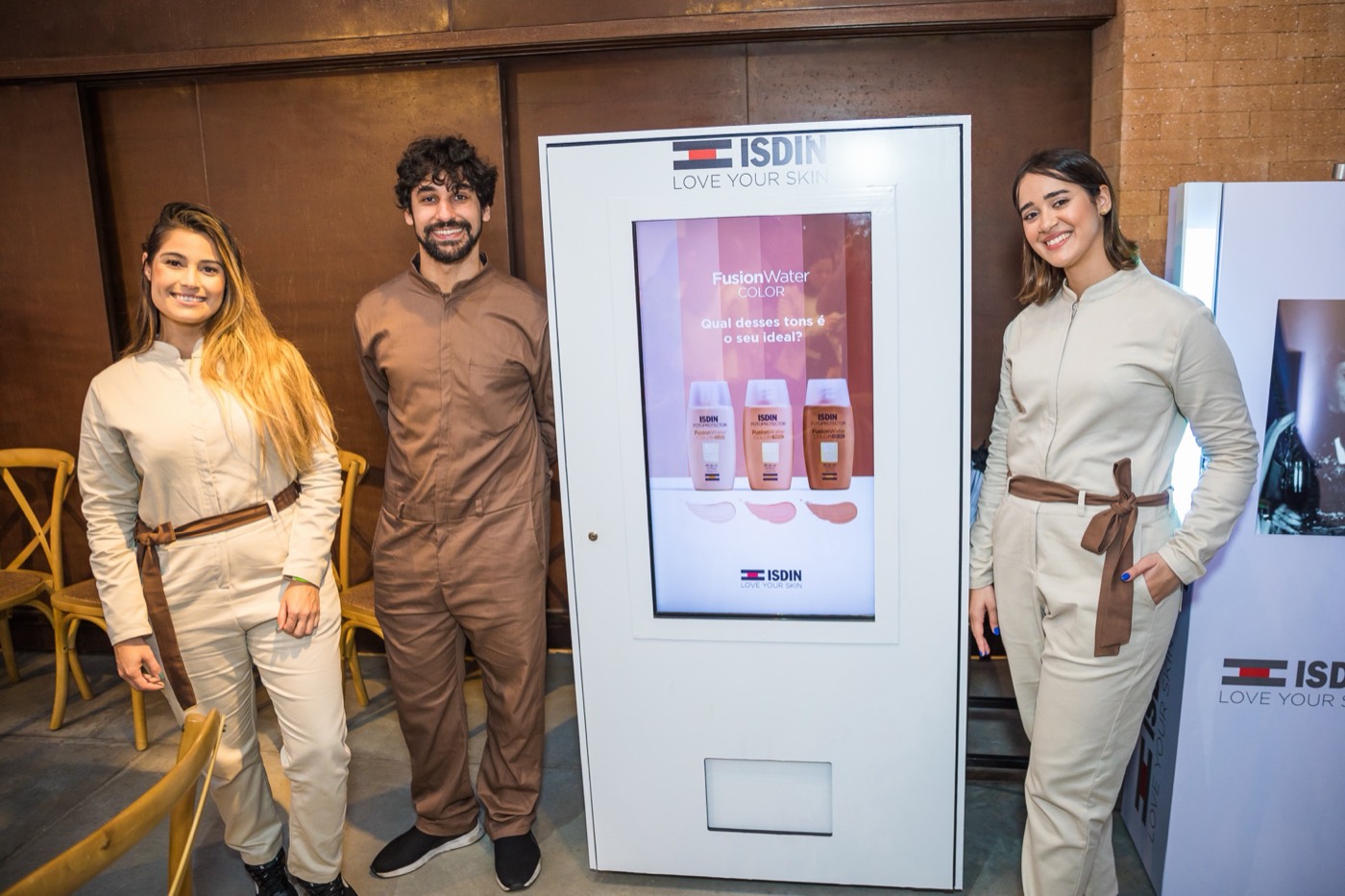 Three people in jumpsuits pose smiling beside a white kiosk with a screen showing product bottles