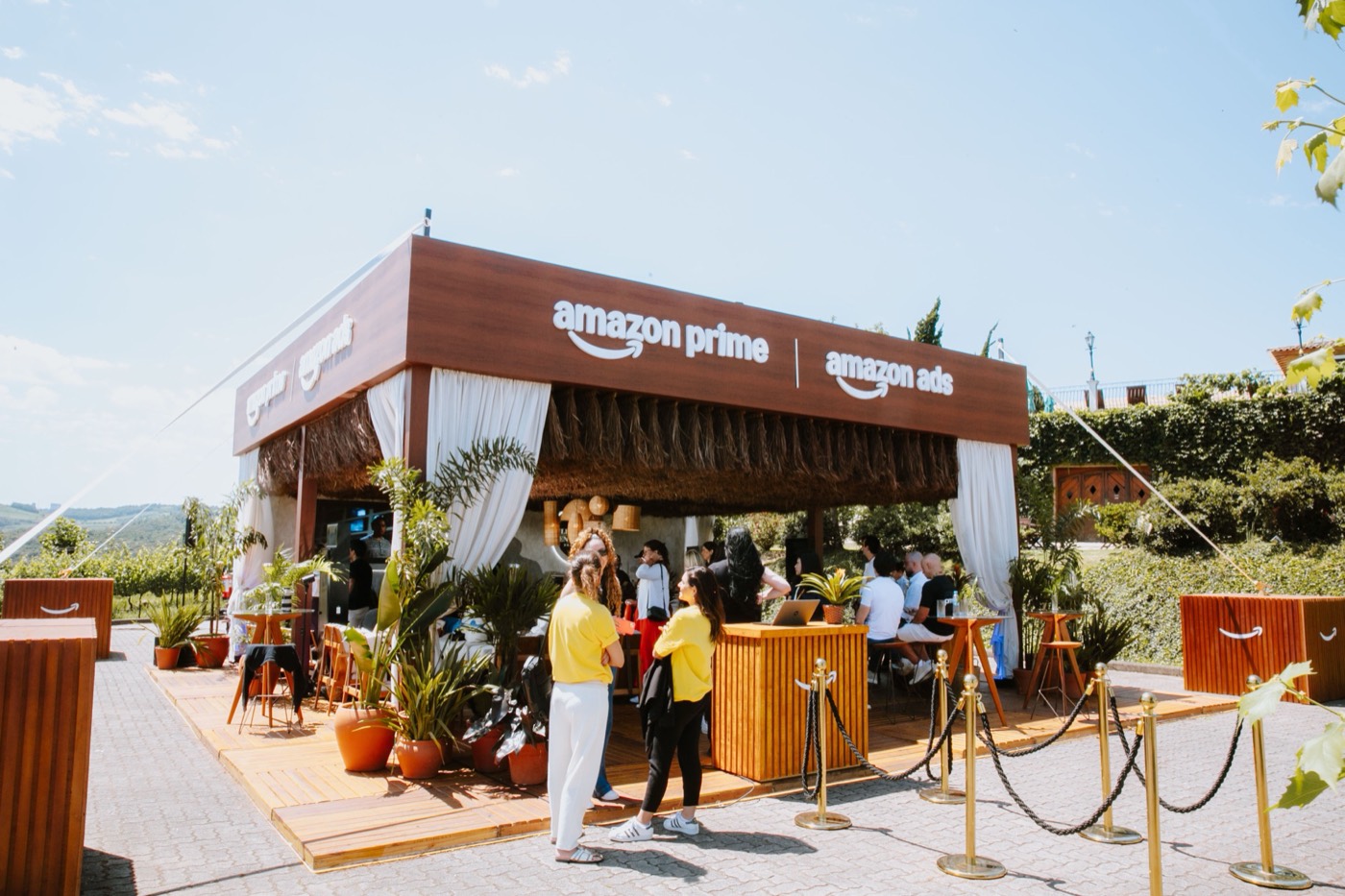 Guests move around an outdoor wooden booth with "amazon prime" and "amazon ads" on the fascia on a sunny day