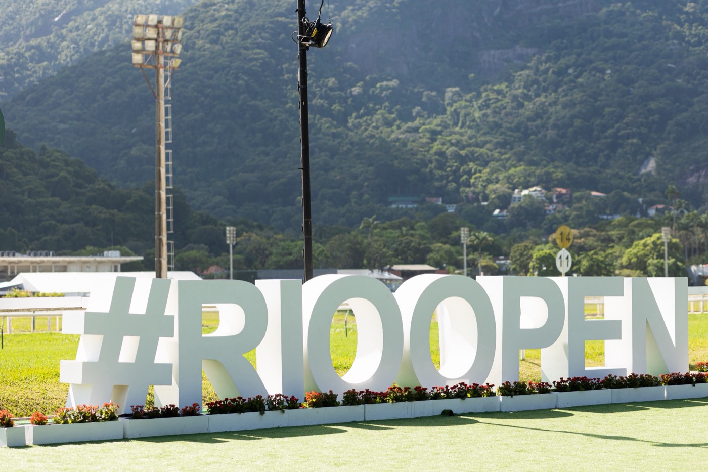 Giant white "#RIOOPEN" sign on a lawn with a flower bed and a wooded hill in the background