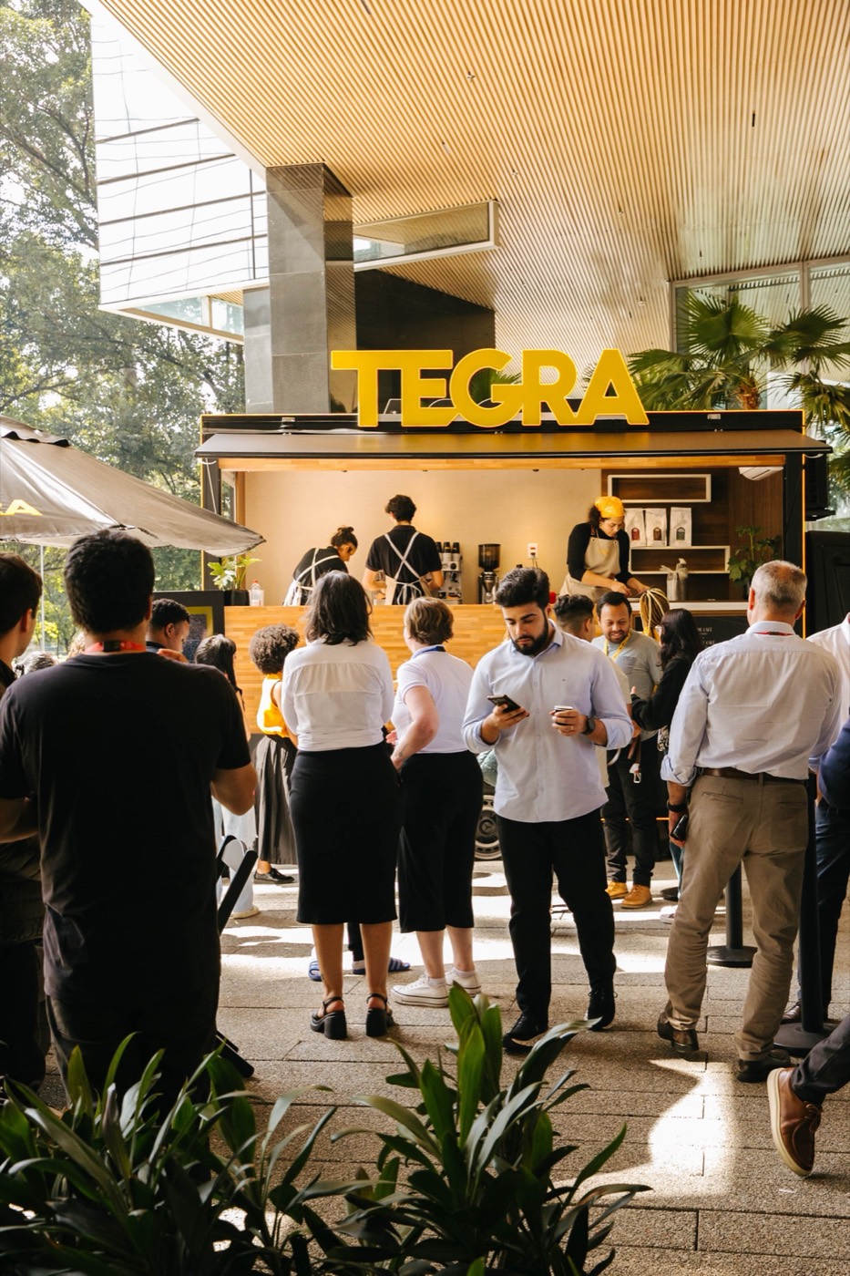 People stand talking and looking at phones by a coffee trailer with a "TEGRA" sign, plants in the foreground