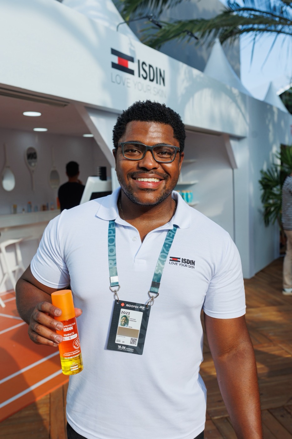 Smiling person in a white polo shirt and badge holds an orange bottle in front of a white "ISDIN" booth
