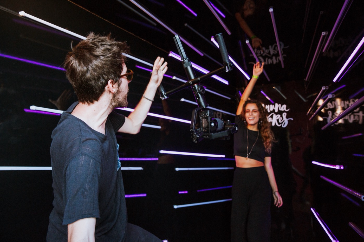 Two people raise their arms by a camera on a stabilizer, in a dark set with purple light bars