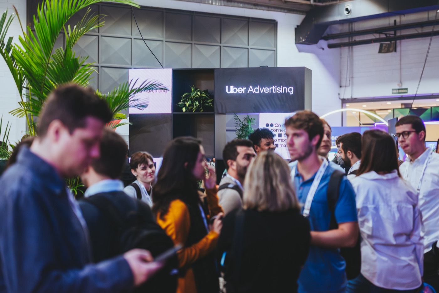 Group of attendees with badges talks in front of a booth with an "Uber Advertising" panel and plants