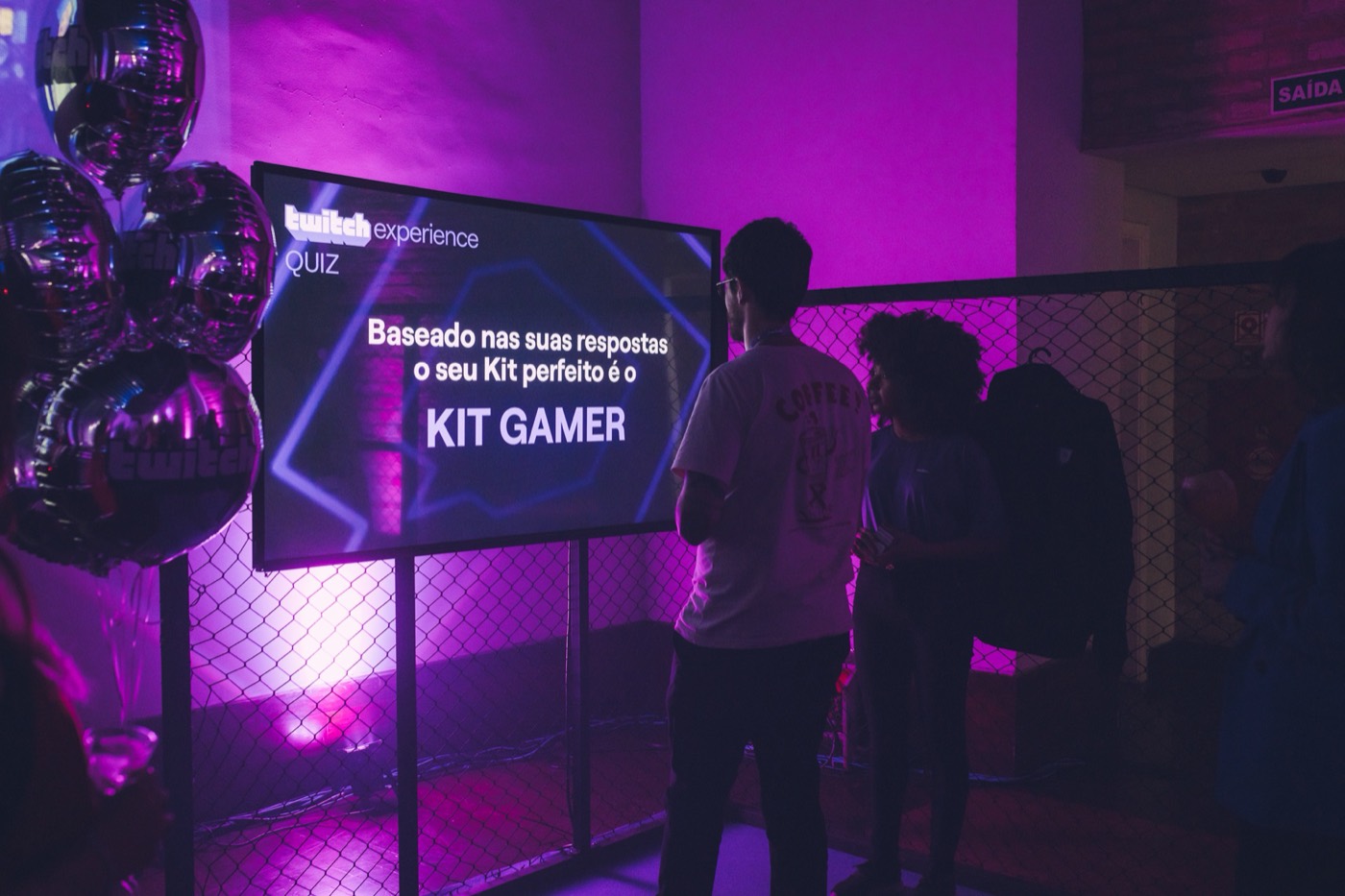 Participants in front of a quiz screen showing the result "KIT GAMER", with metallic balloons and purple light