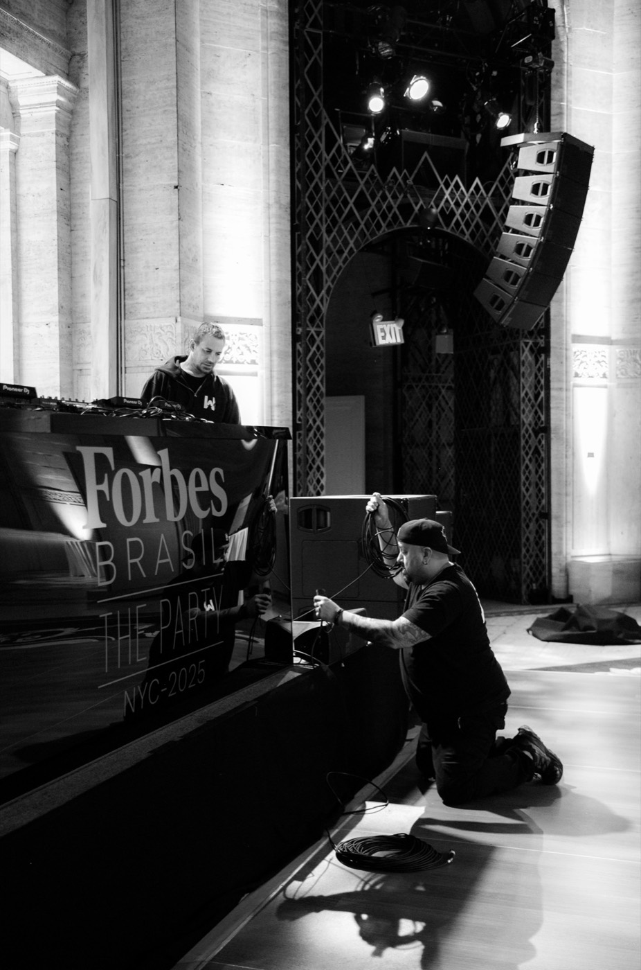 Black and white backstage, with a person kneeling to arrange cables by the stage and another at the sound booth
