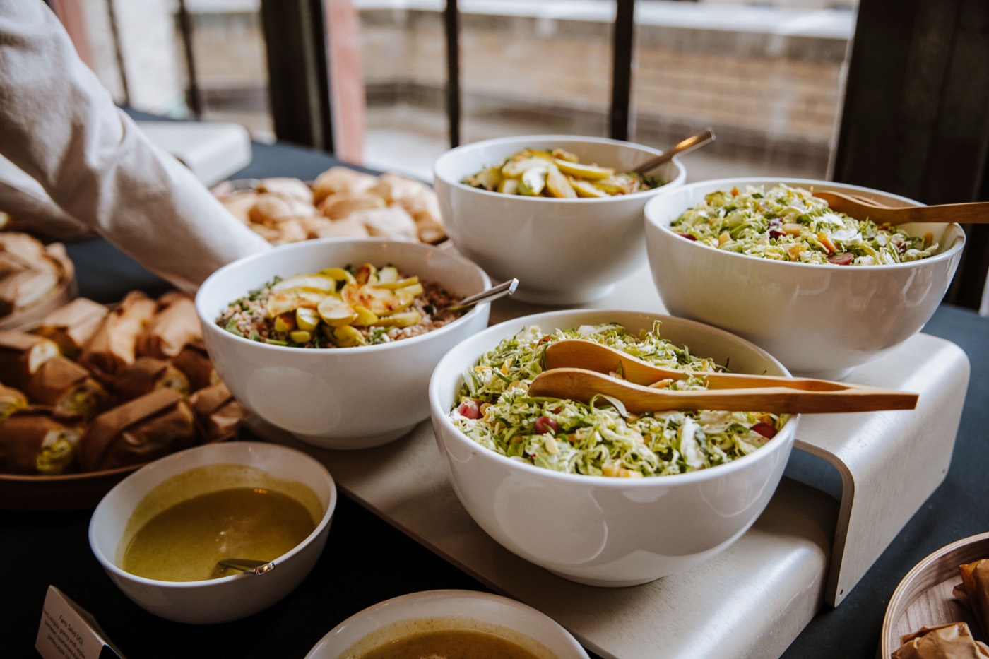 Buffet with white bowls of salads, wooden tongs and bread, with a person's arm serving in the background