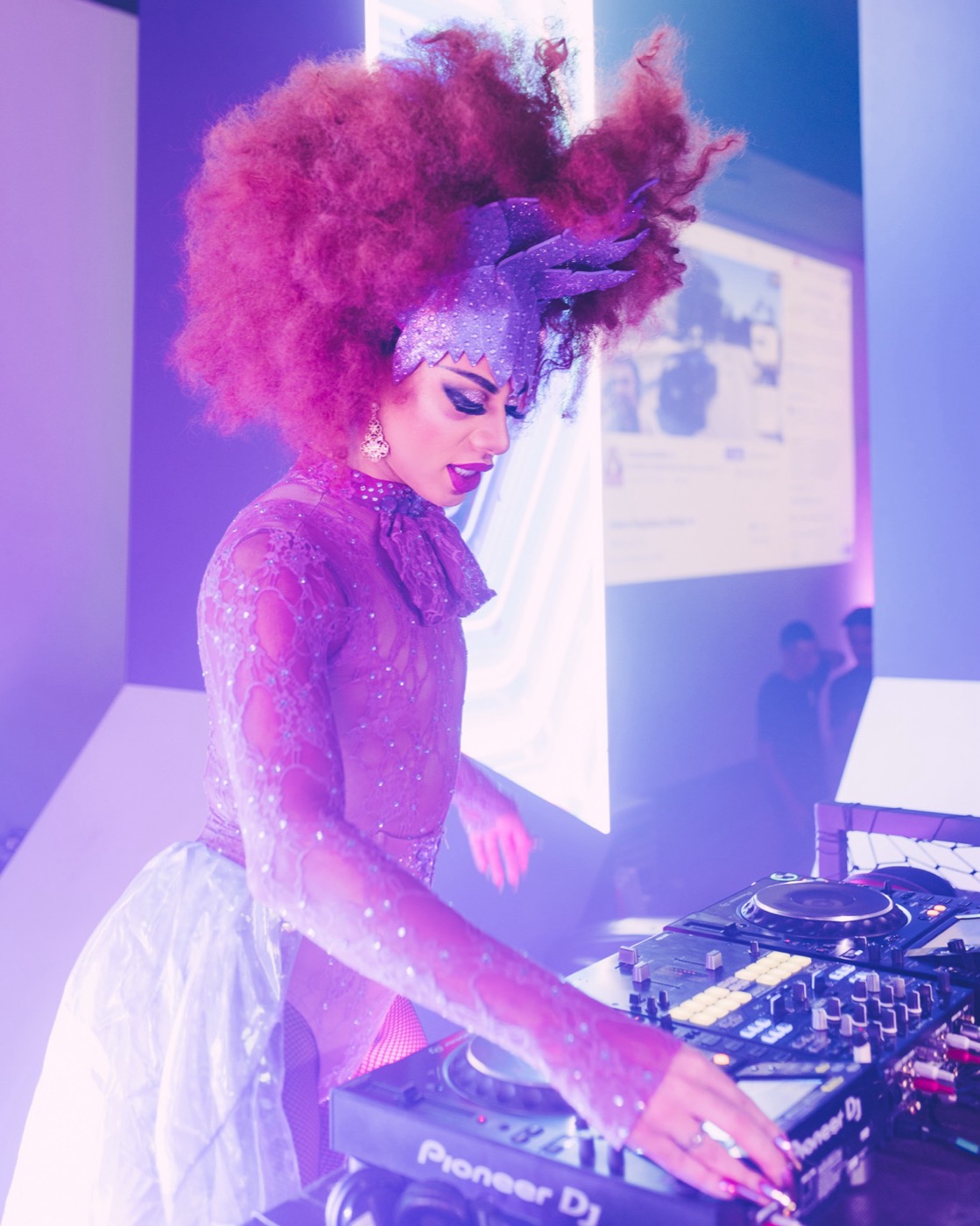 DJ with voluminous red hair and a sparkly outfit works the mixing desk