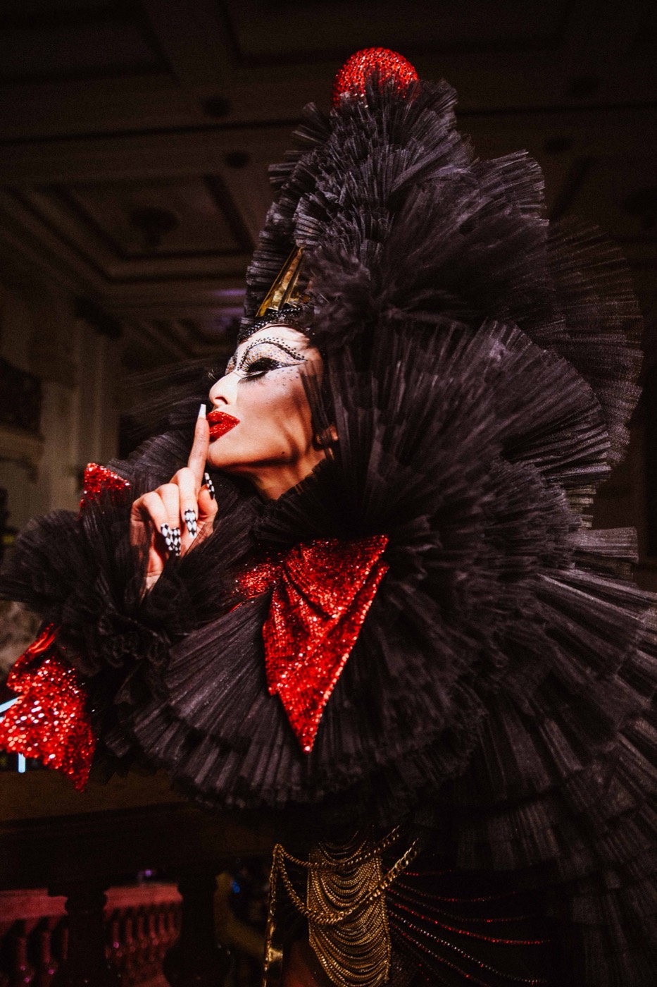 Profile of a person in theatrical makeup, a black tulle costume and red bow, with a finger to their lips