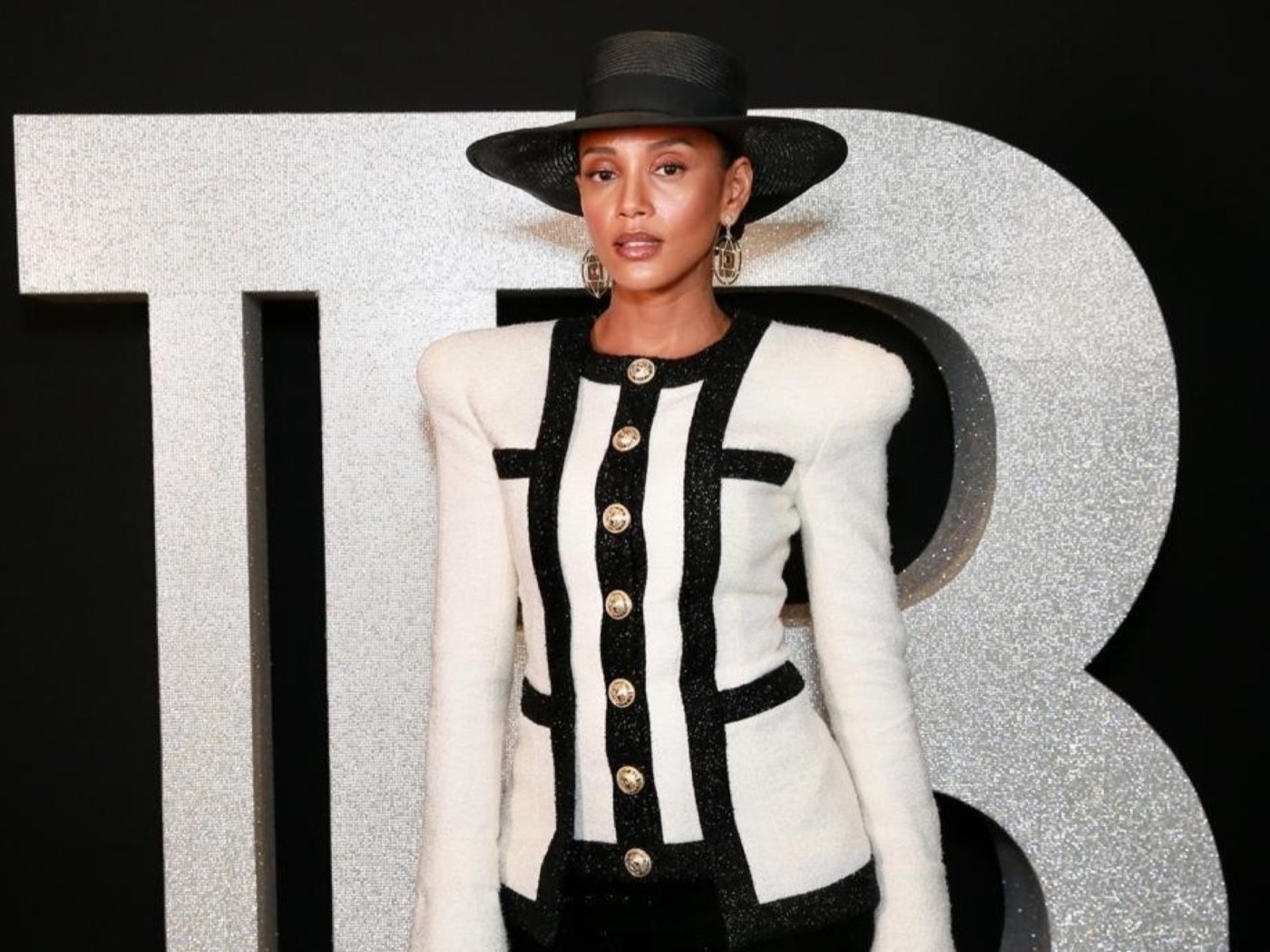 Person in a black hat and white blazer with black stripes and gold buttons before a glittery silver letter "B"