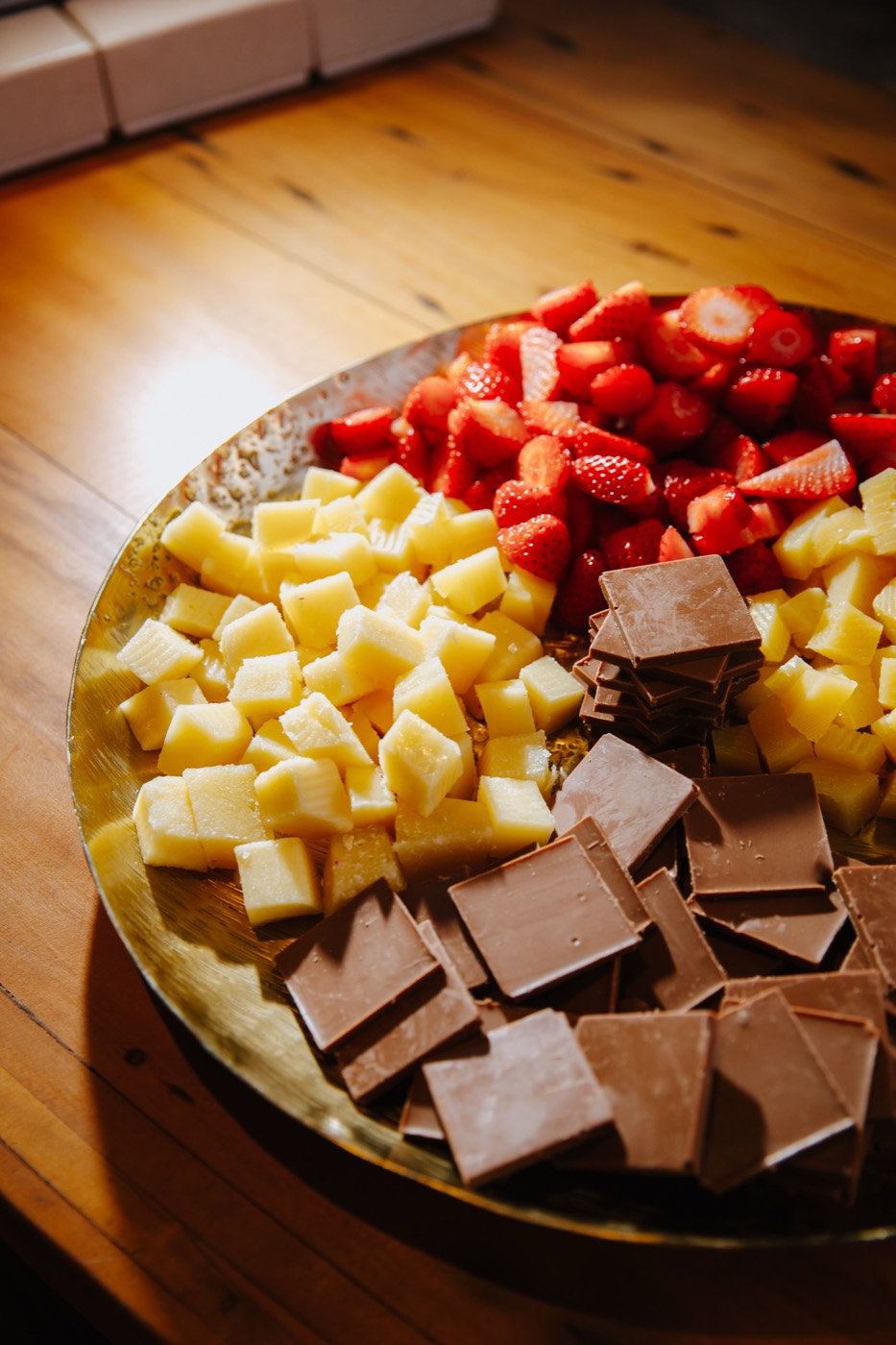 Gold tray with chopped strawberries, yellow cubes and chocolate squares on a wooden table