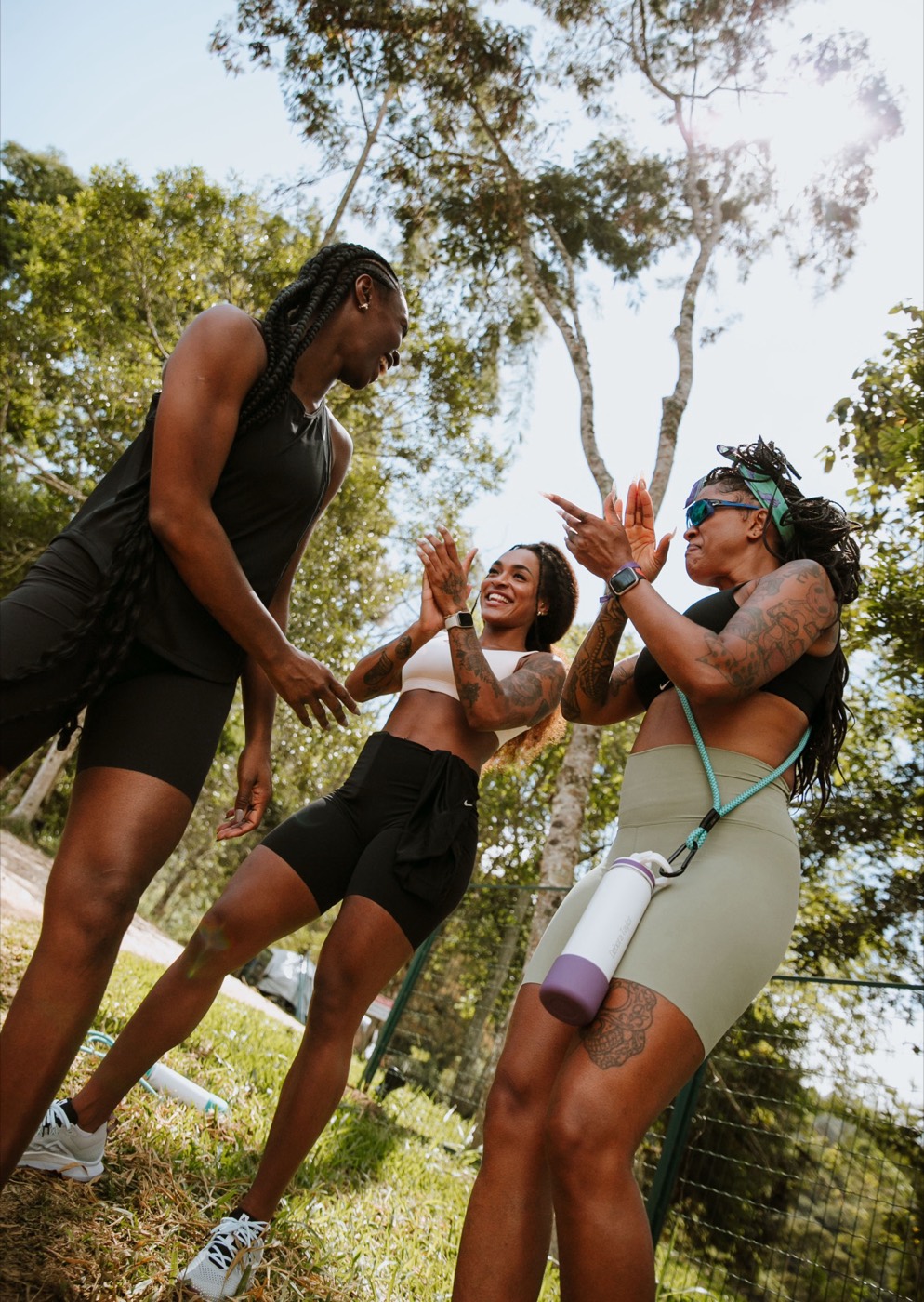Three people in workout clothes talk and gesture outdoors under trees, seen from below