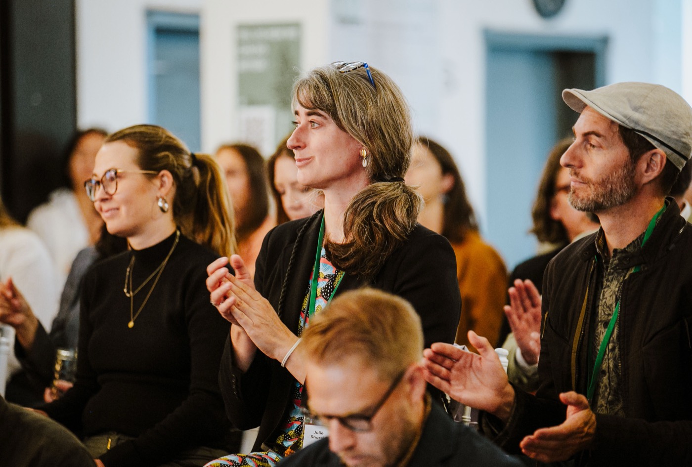 Seated audience applauds and smiles during a presentation, with participants wearing green lanyard badges
