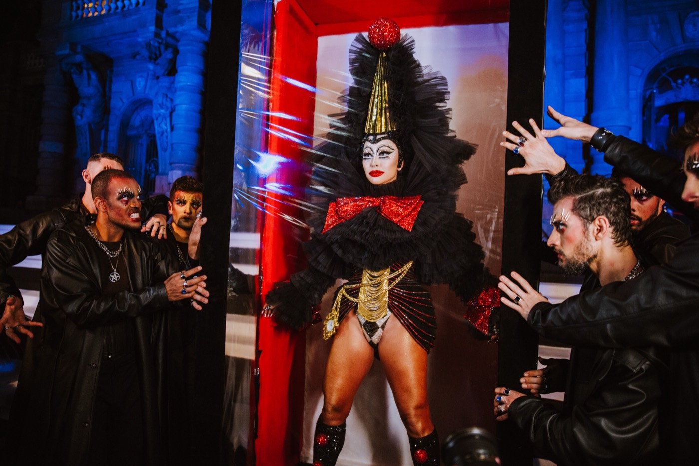 Person in a black costume and pointed hat inside a red booth, with other people's hands reaching toward them