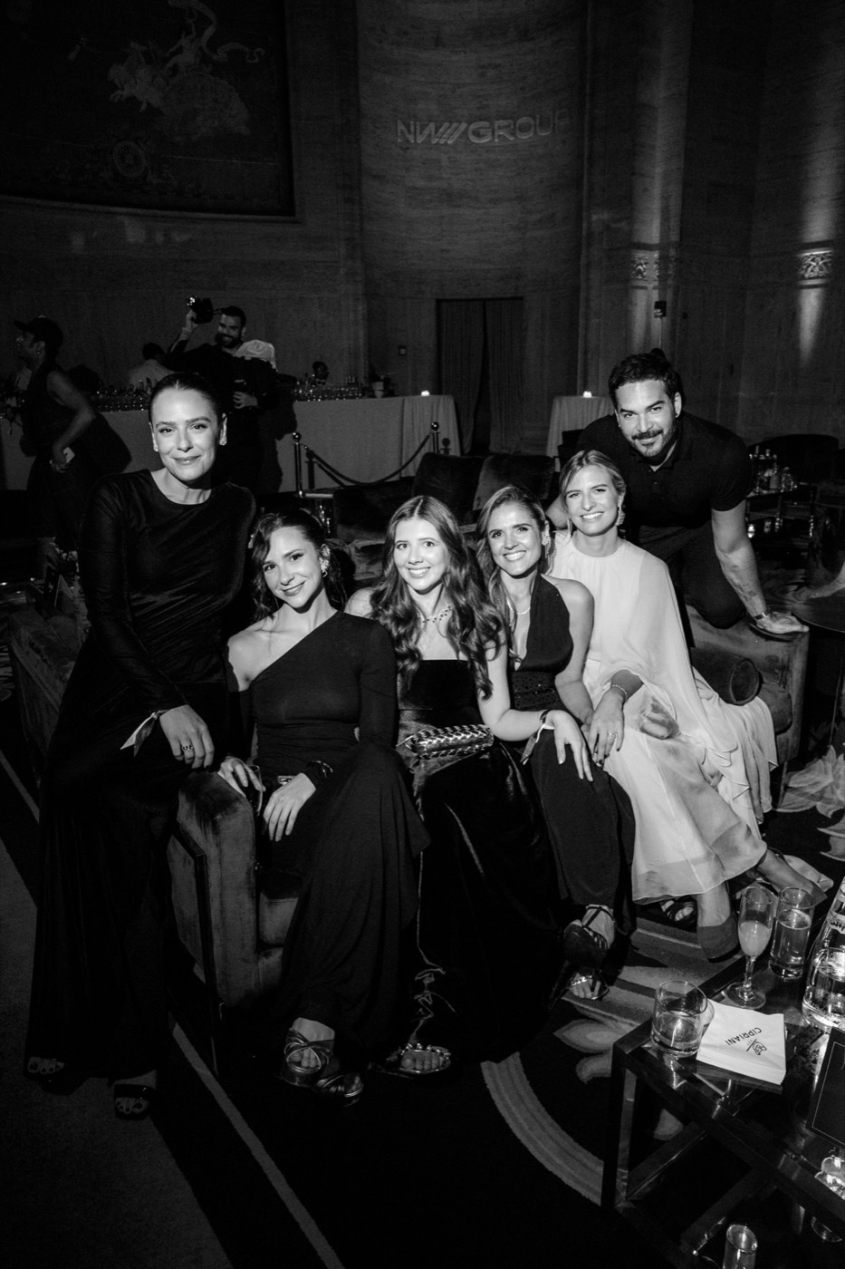 Six guests in evening wear pose smiling on lounge sofas, in black and white