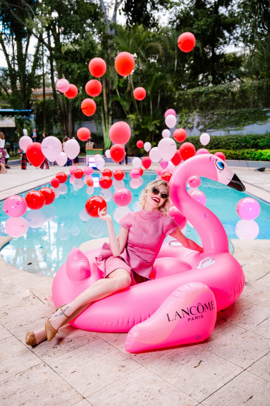 Person in a pink dress and sunglasses poses on a giant "LANCÔME" flamingo float by a pool with balloons