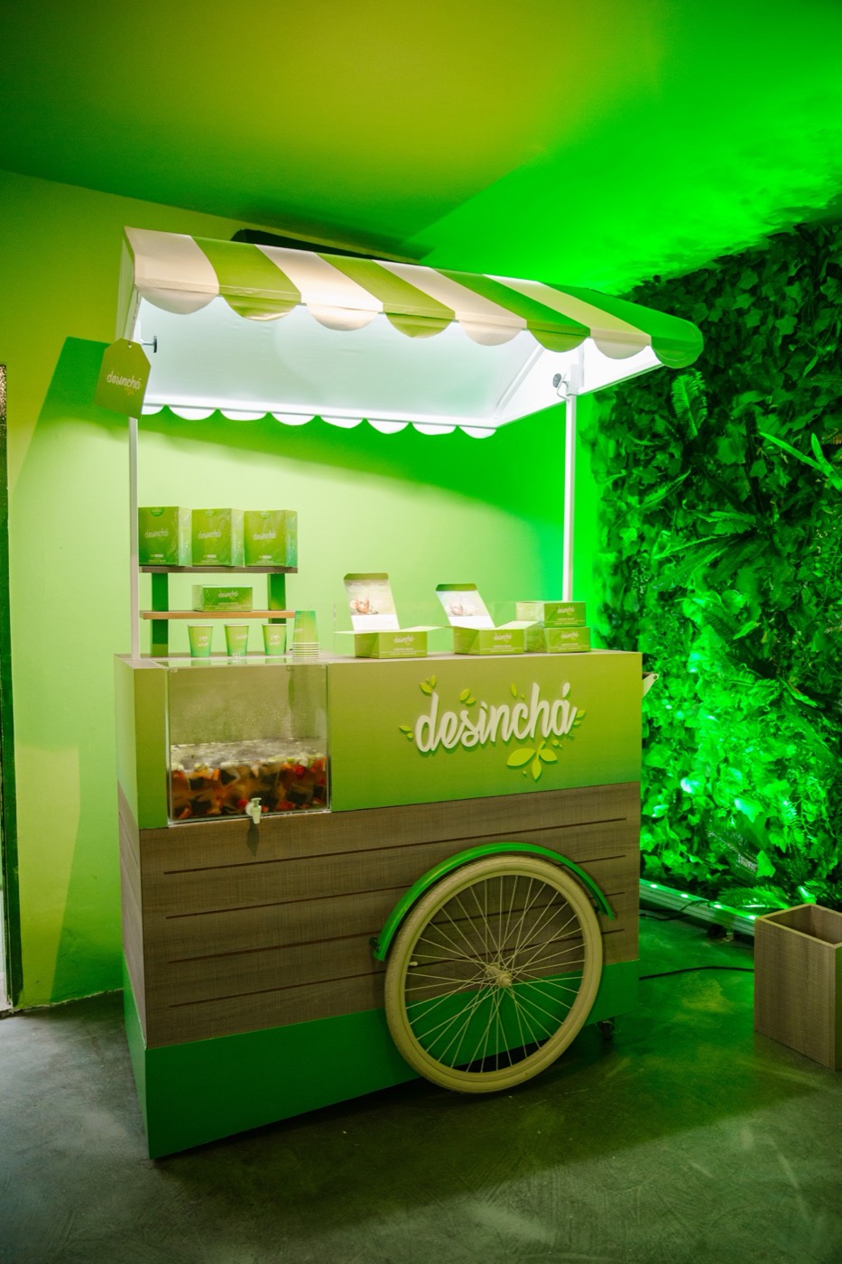 Green cart with a striped awning, product boxes and "desinchá" lettering, next to a foliage wall