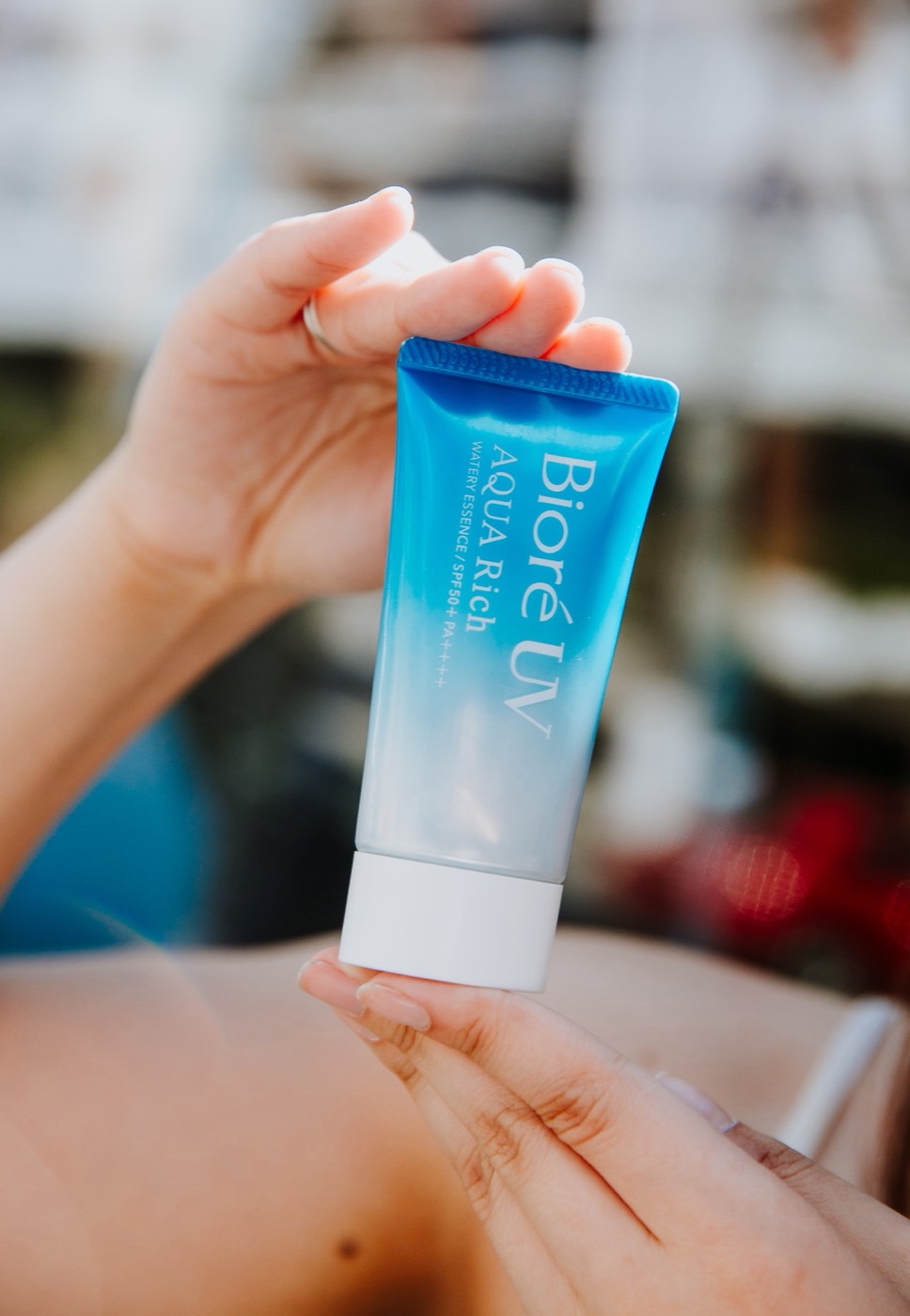 Hands hold a blue "Bioré UV AQUA Rich" tube in close-up, with a blurred background