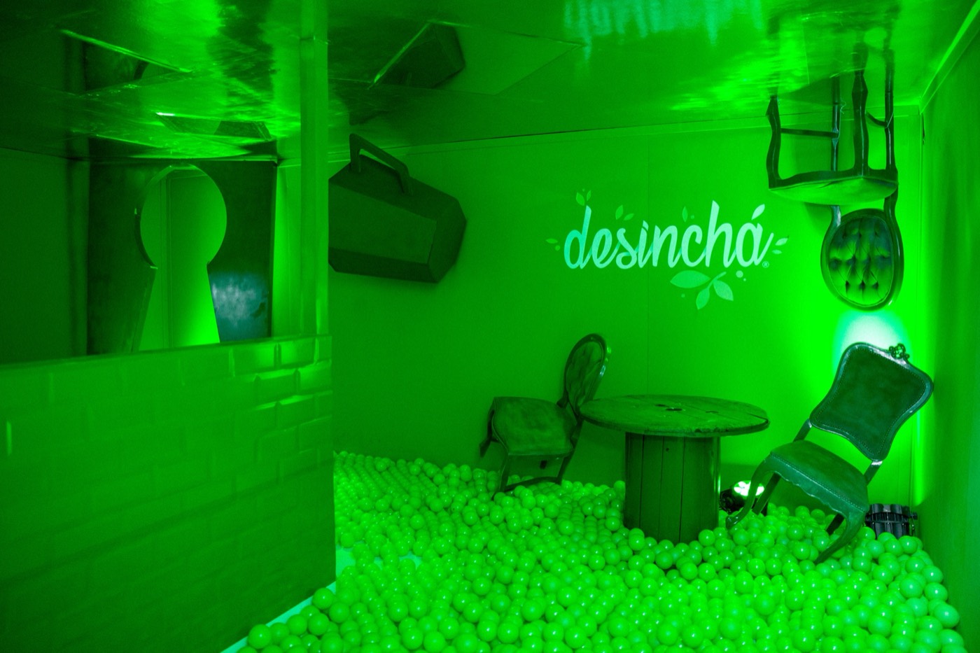 Green-lit room with a ball pit, a table and chairs, furniture fixed to the ceiling and a "desinchá" projection