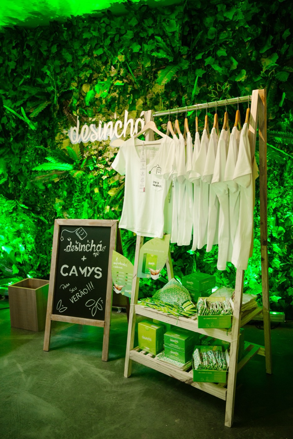 Clothing rack with white T-shirts and product boxes in front of a foliage wall, with a chalk-written board