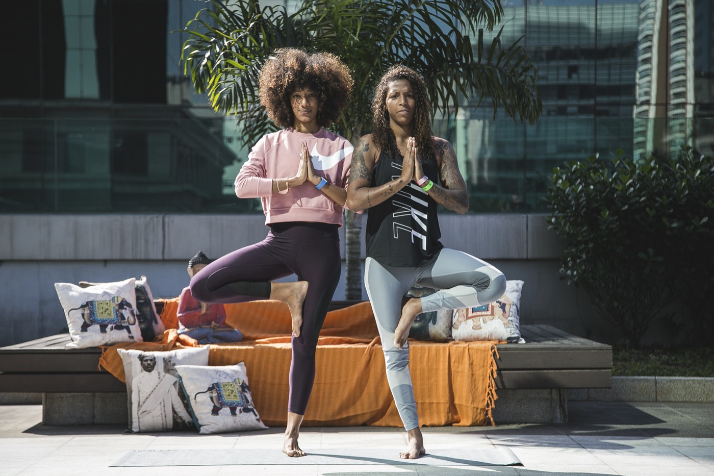 Two people standing in tree pose with hands together, in front of a bench with an orange throw and cushions