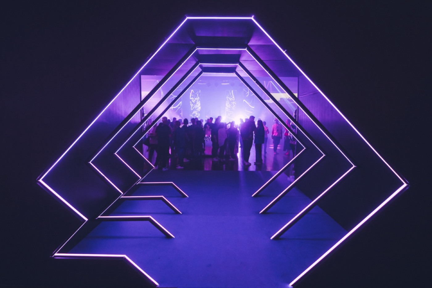 Tunnel of purple neon light frames leads to a hall with the crowd in silhouette