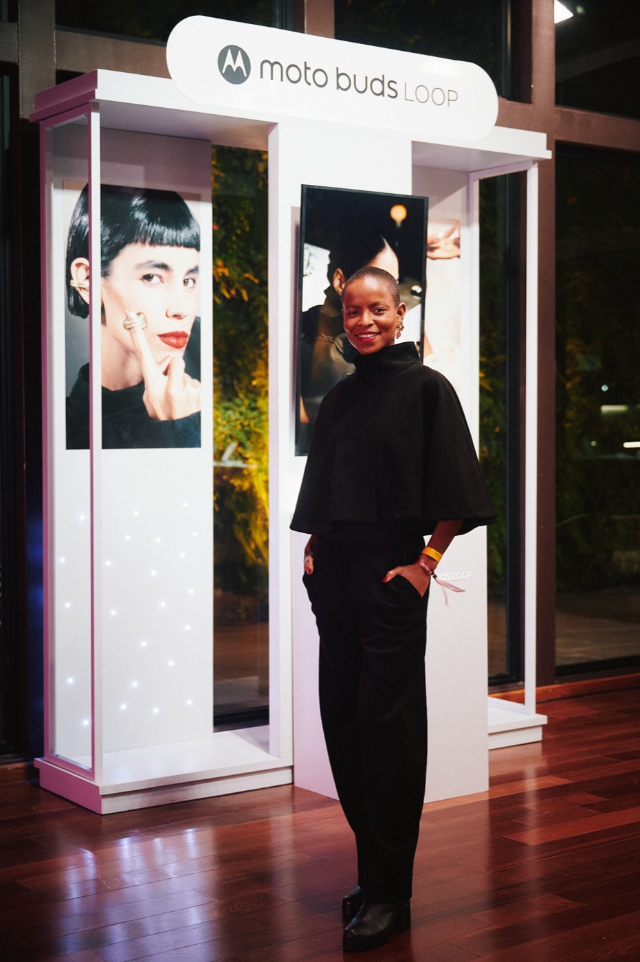 Person in black clothing poses smiling next to a white display case with a "moto buds LOOP" sign