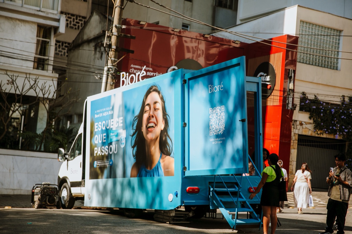 Blue truck with an "ESQUECE QUE PASSOU" panel and "Bioré" lettering parked on the street, with people by its stairs