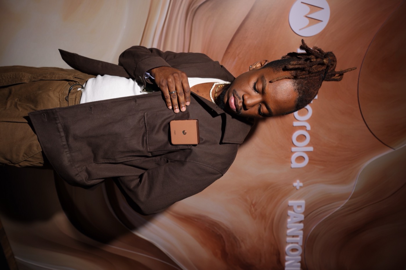 Person in a brown jacket touches a flip phone tucked into the chest pocket, before a "motorola + PANTONE" panel