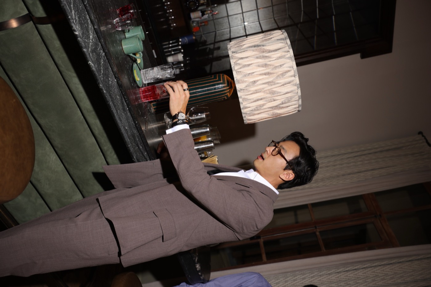 Person in a suit and glasses handles a glass of red drink at a bar counter, beside a table lamp