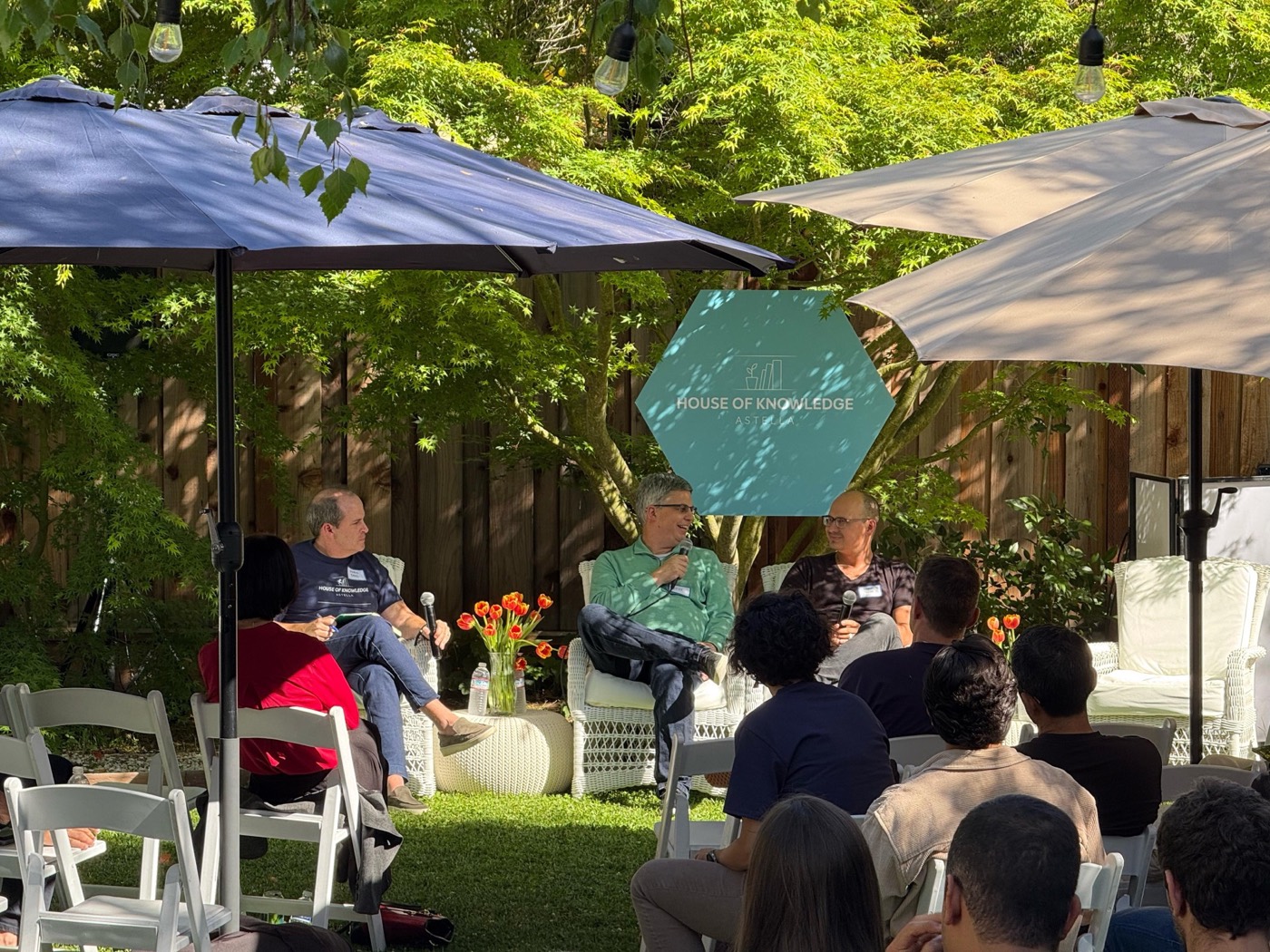Garden talk with three people seated under umbrellas in front of a hexagonal "HOUSE OF KNOWLEDGE" sign, audience in foreground
