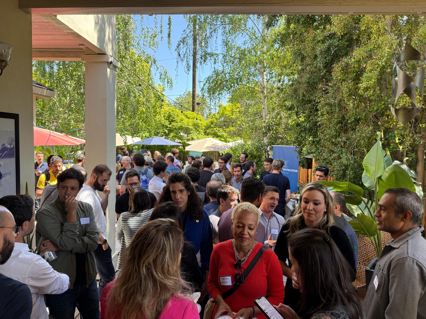 Guests with name tags stand chatting in a crowded, tree-lined garden, seen from a porch