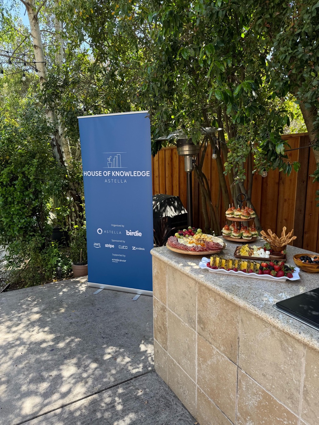 Blue "HOUSE OF KNOWLEDGE ASTELLA" banner next to a stone counter with charcuterie boards, fruit and snacks