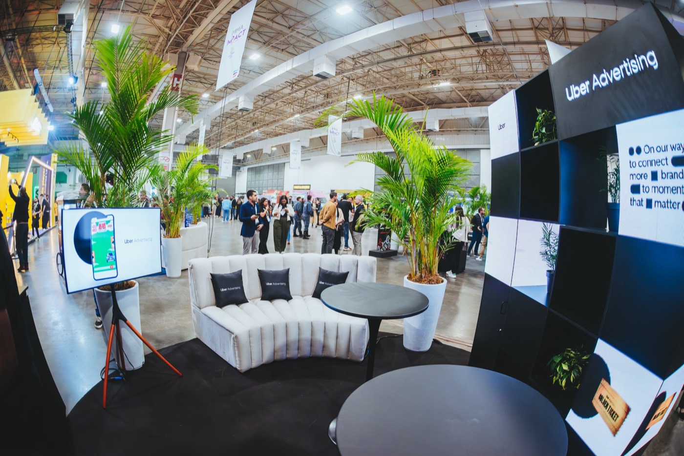 Booth lounge with a curved white sofa, black tables, palm trees and an "Uber Advertising" panel, in a large hall