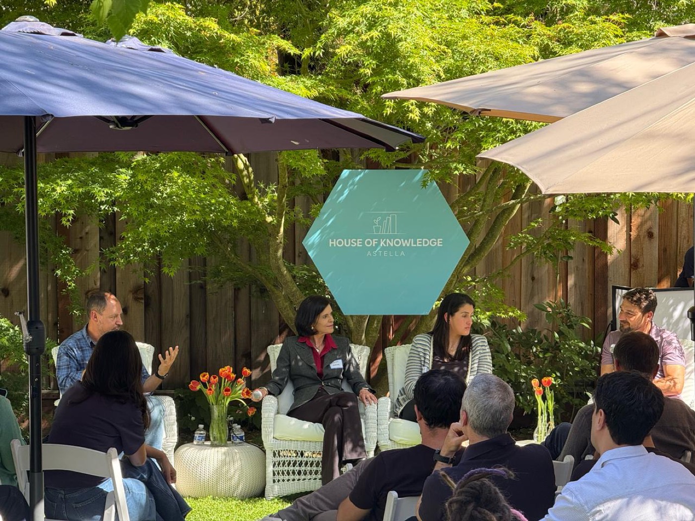 Four people seated in white armchairs talk in a garden in front of a "HOUSE OF KNOWLEDGE" sign