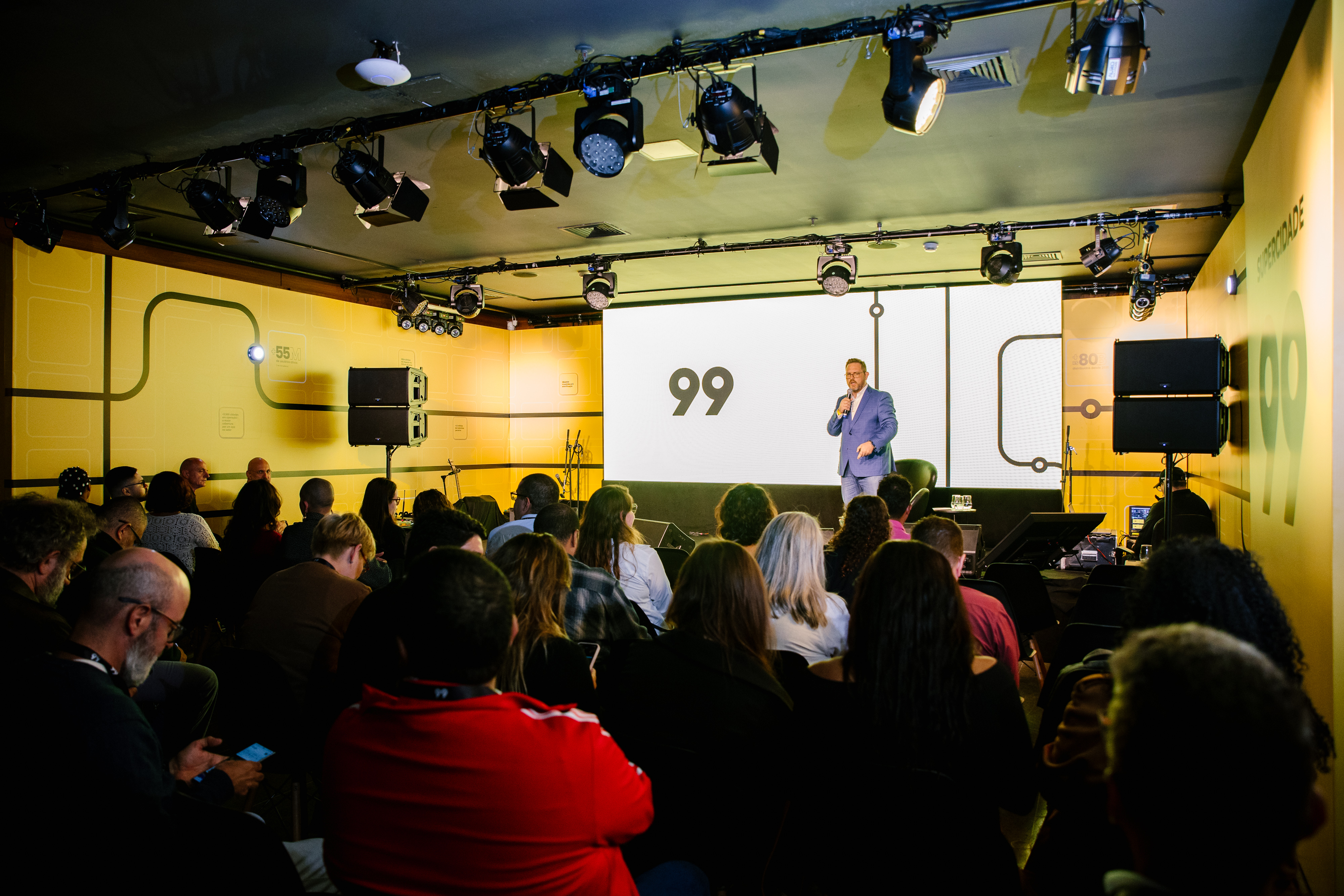 Seated audience watches a person with a microphone on stage in front of a screen showing "99"