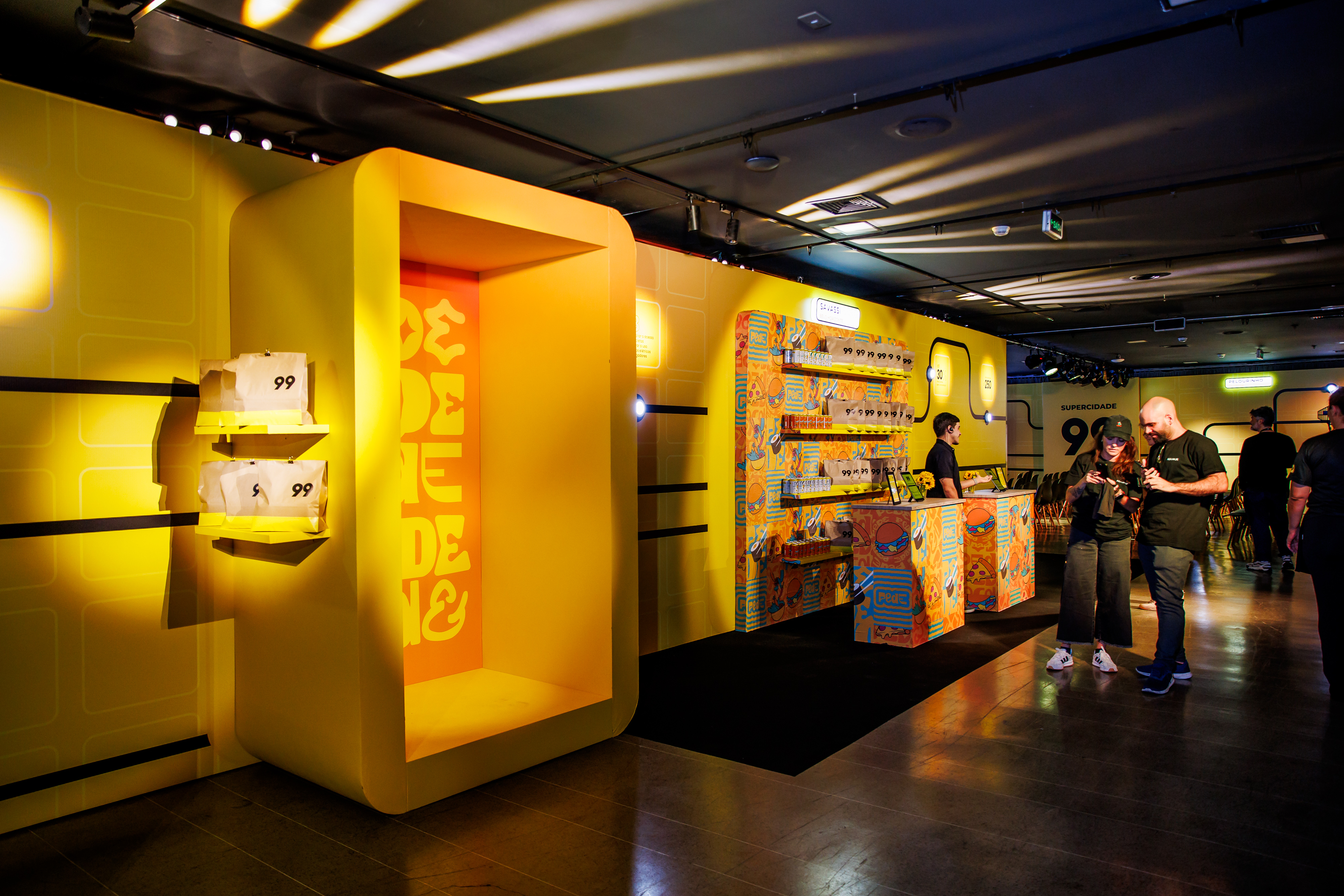 Hallway with yellow scenography, a lit niche, a display of "99" bags and people chatting in the background