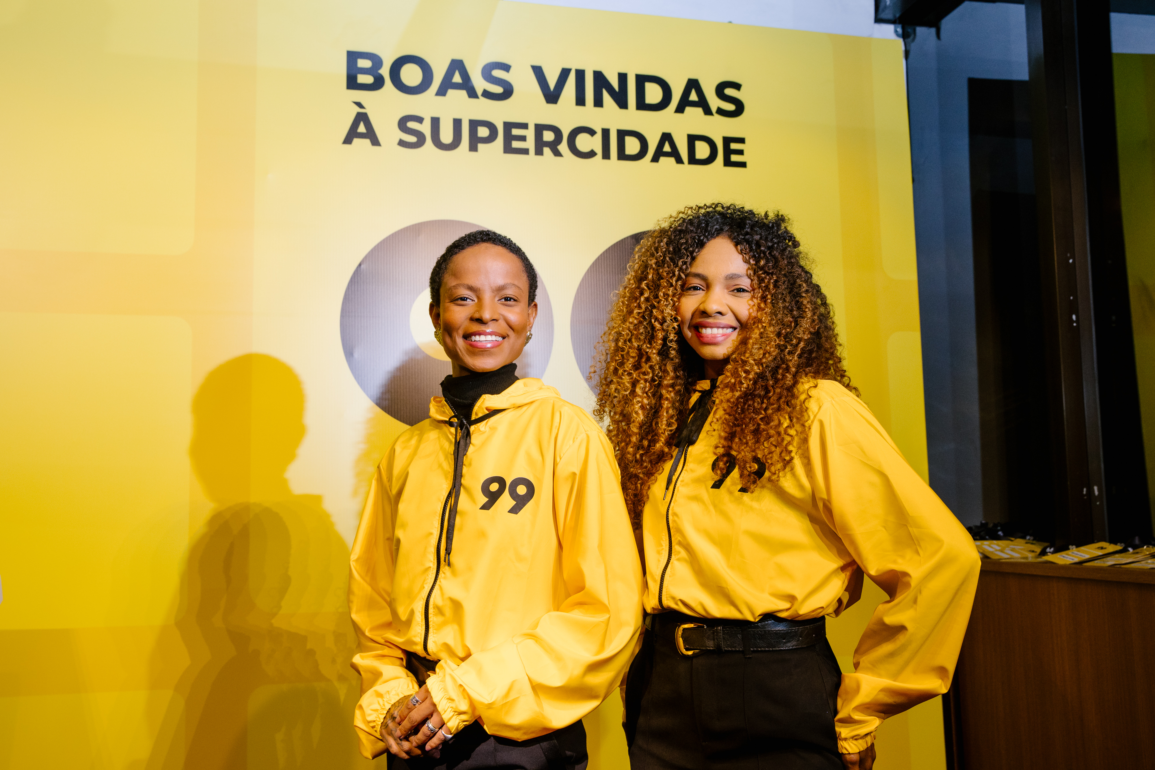 Two people in yellow "99" jackets smile in front of a "BOAS VINDAS À SUPERCIDADE" panel